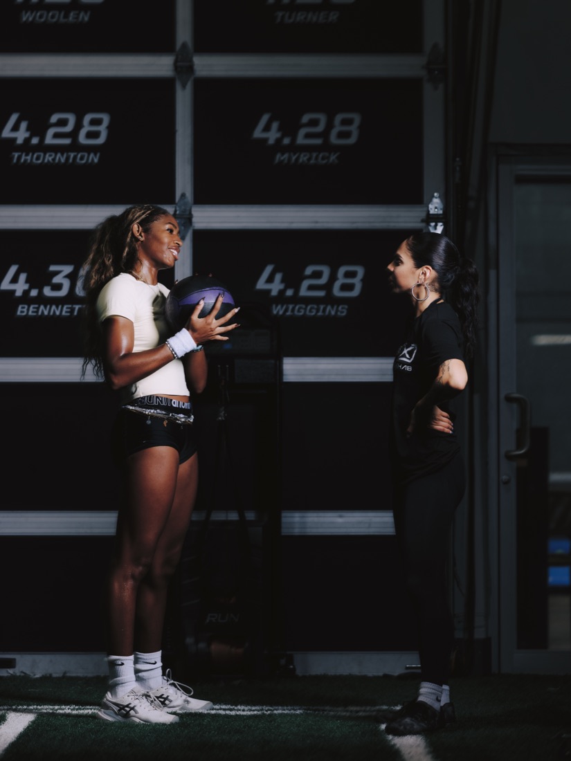 An athlete holding a medicine ball in front of the 40-yard times wall, with Nicole beside her.